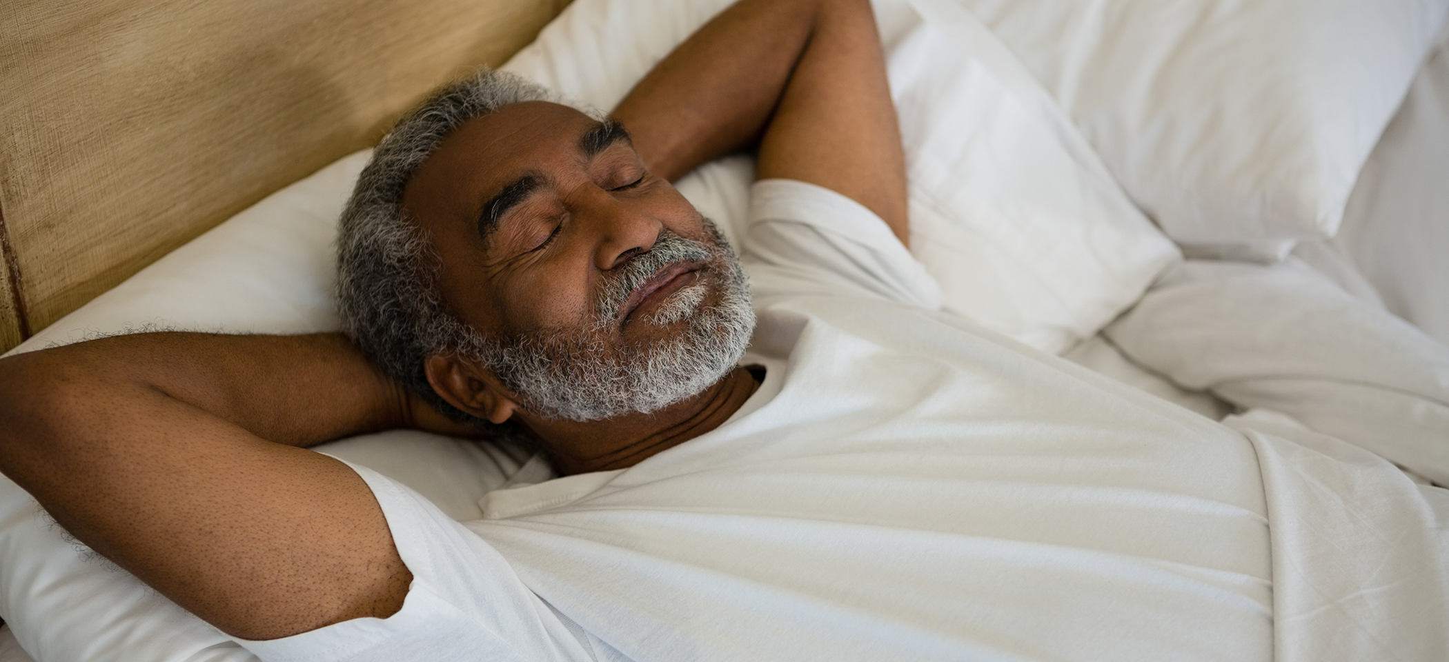 Bearded man sleeping in bed with hands behind head